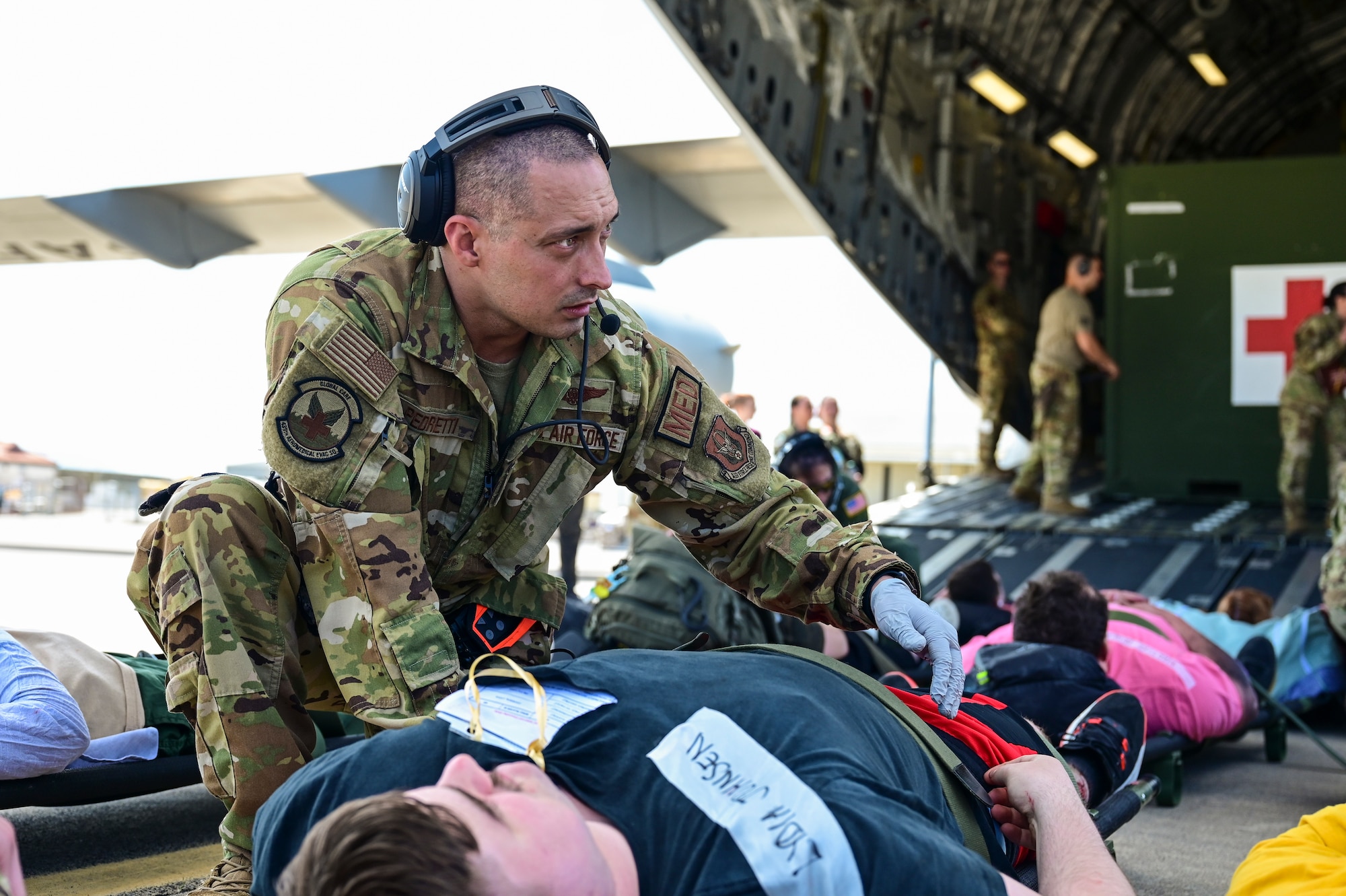 An Airman in a camo flight suit kneels on the ground at the back of an open C-17 Globemaster III as he attends to a patient on a stretcher.