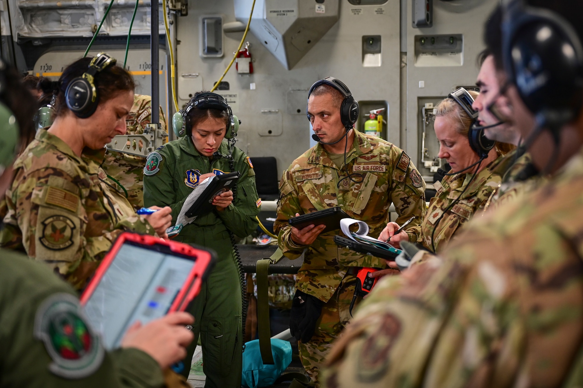 Men and women in the Air Force, dressed in flight suits and camo all wearing headsets, review notes on iPads in their hands while standing in a circle.
