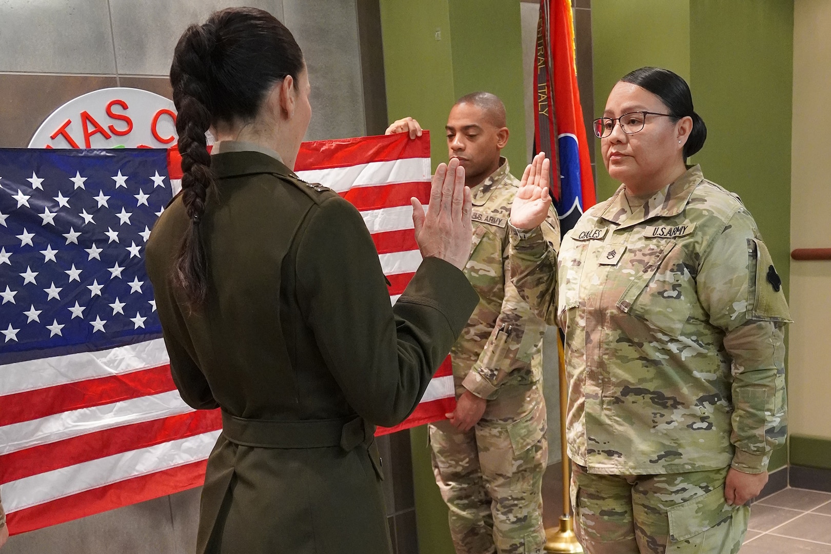 Two women in military uniform stand before each other with their right hands raised. A man in uniform stands in the background holding the American flag.