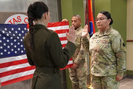 Two women in military uniform stand before each other with their right hands raised. A man in uniform stands in the background holding the American flag.