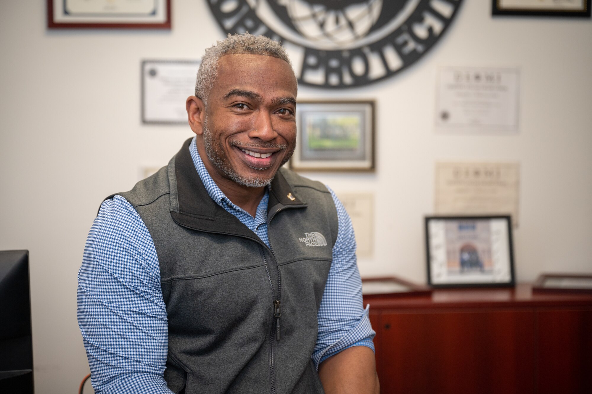 A man poses in his office and smiles at the camera. Behind him is a steel cut out of a phoenix raven unit patch;