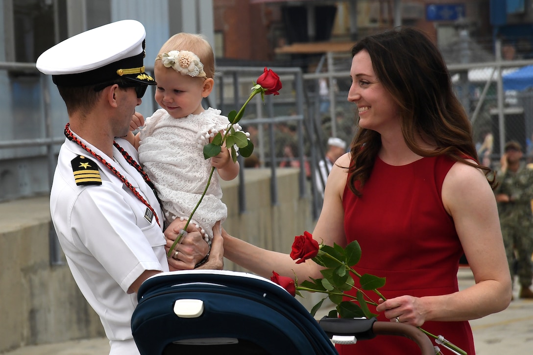 A sailor in uniform holds a child as his wife smiles. Both the infant and the woman are holding roses.