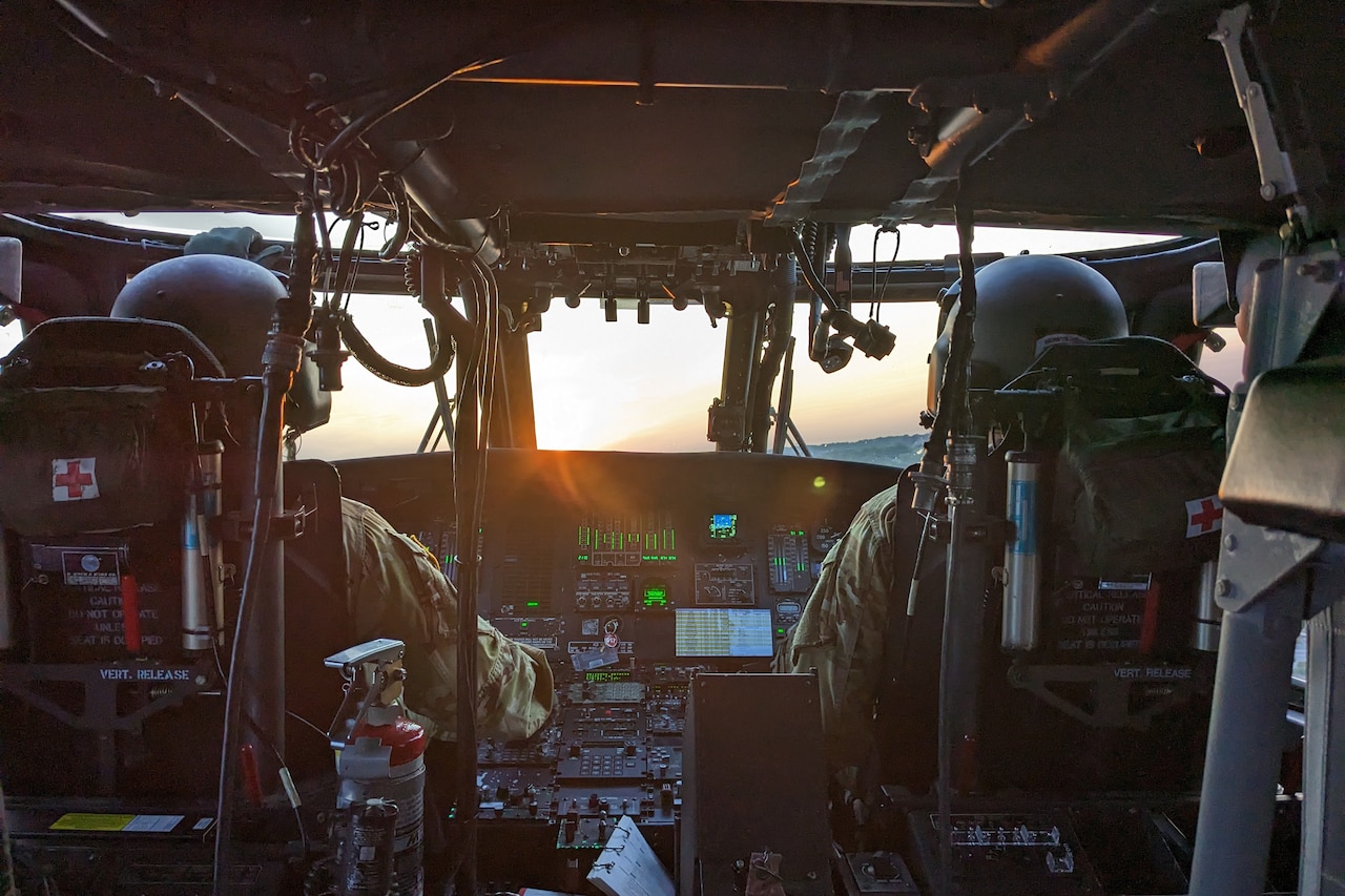 A view of two pilots flying from inside the cockpit of a helicopter at sunset.