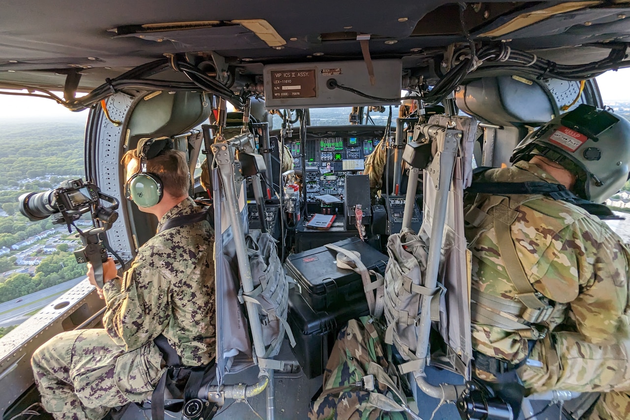 A man holding a video camera points it out the left side of a flying helicopter. Another man sits behind him as two men pilot the aircraft.