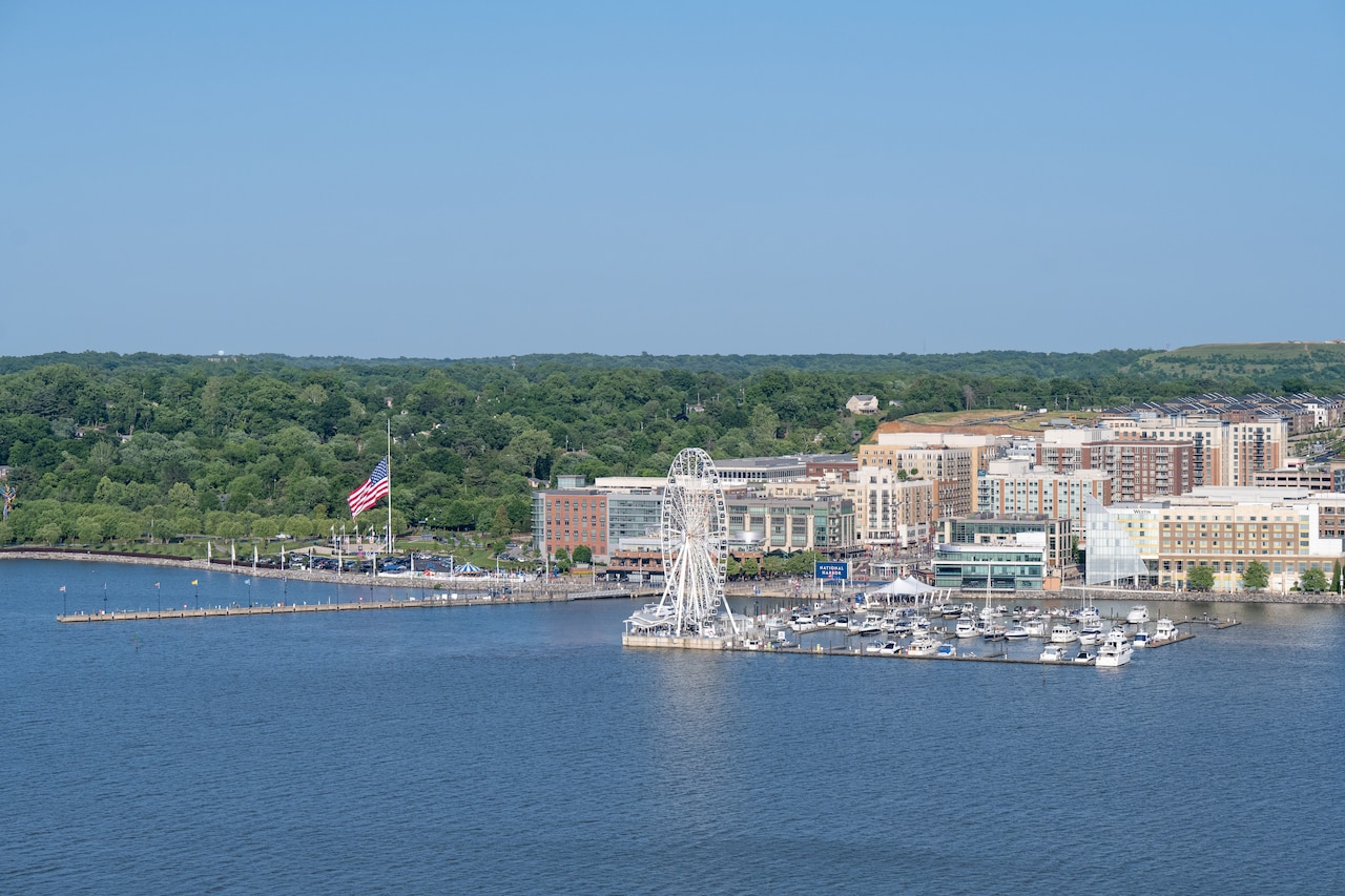 An aerial view of a ferris wheel on a dock beside several boat slips. Behind it on the coast are large buildings.