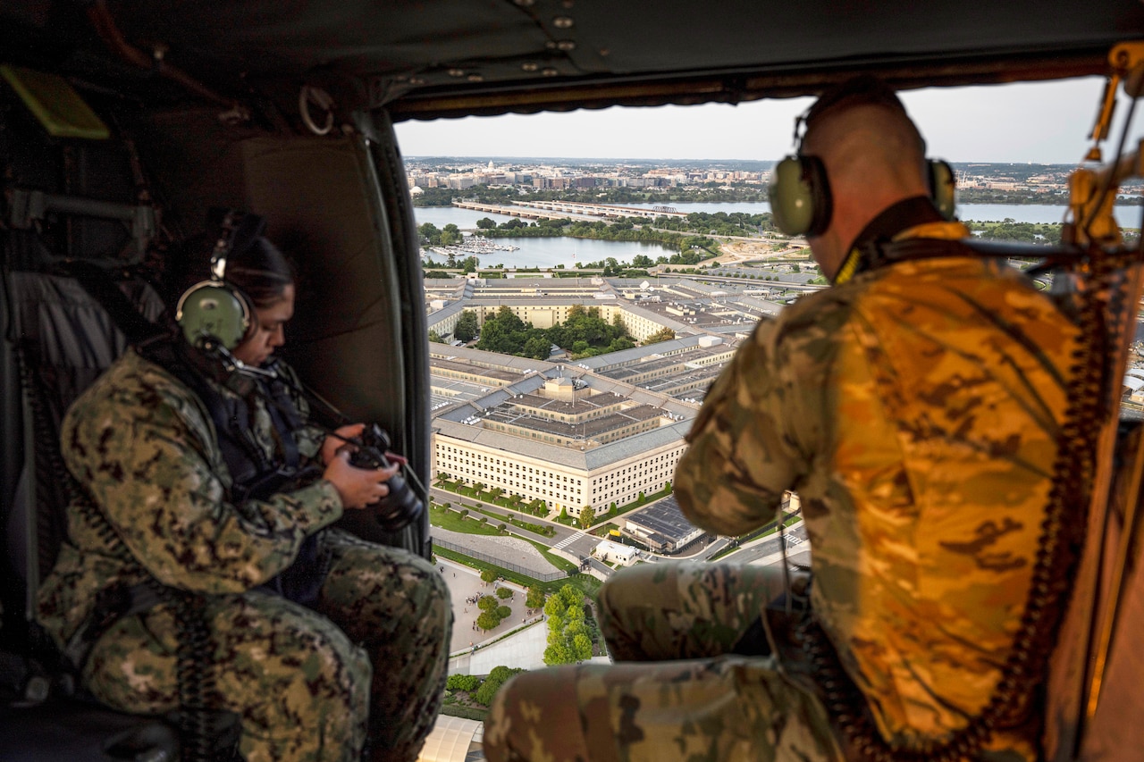 Two photographers strapped into a helicopter aim their cameras toward a building.