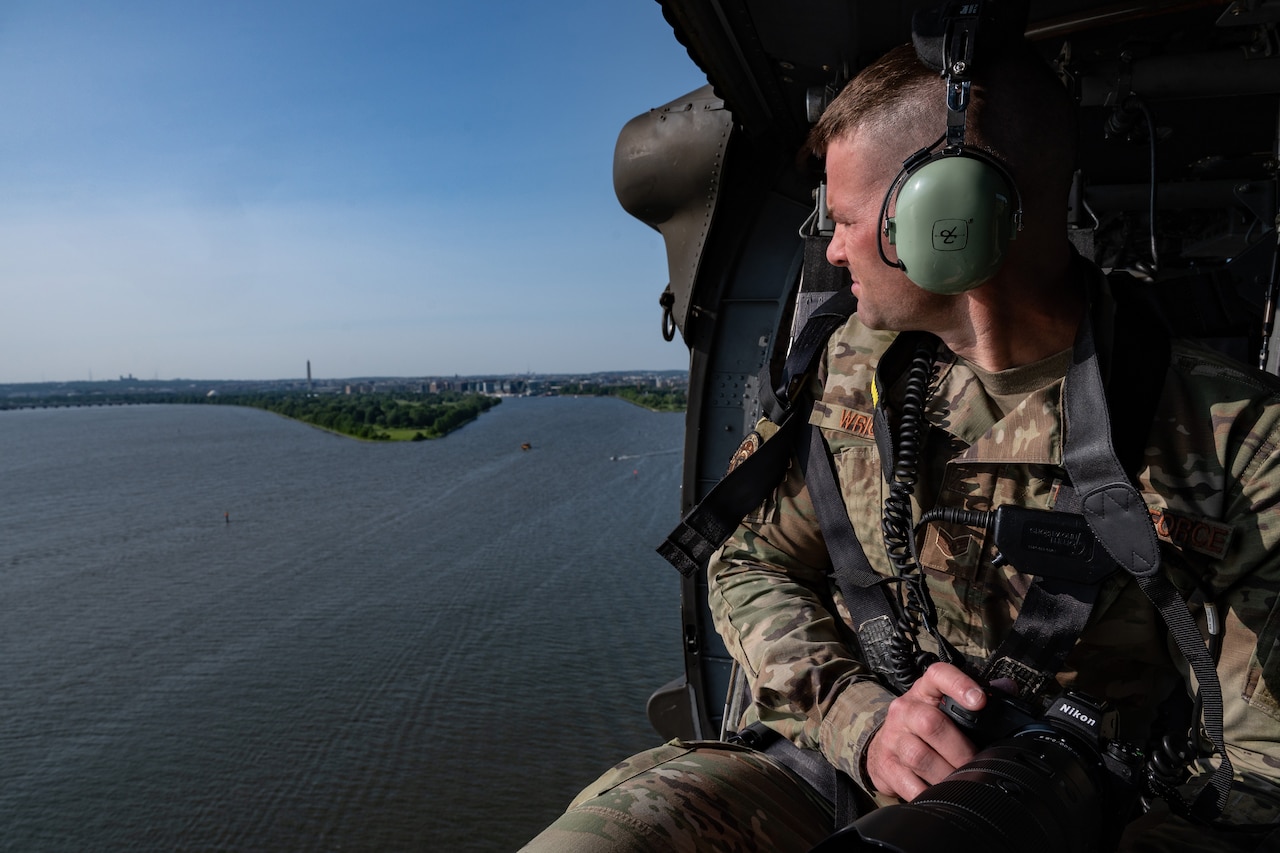 A man holding a camera looks out the side of a helicopter.