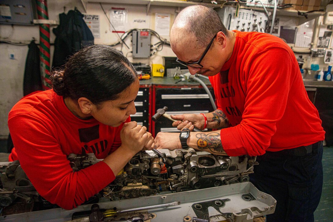 Two sailors work together on a mechanical part while on a ship at sea.