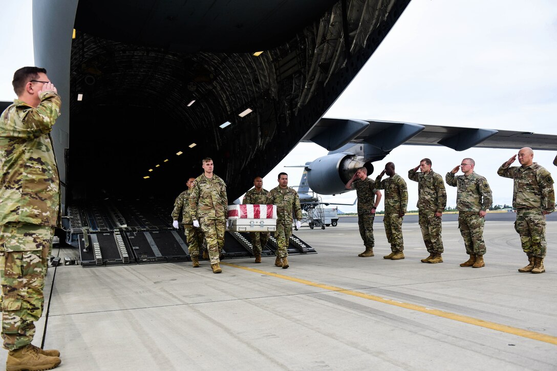 A line of service members salute on either side of the back of an aircraft as others carry a flag-draped coffin.