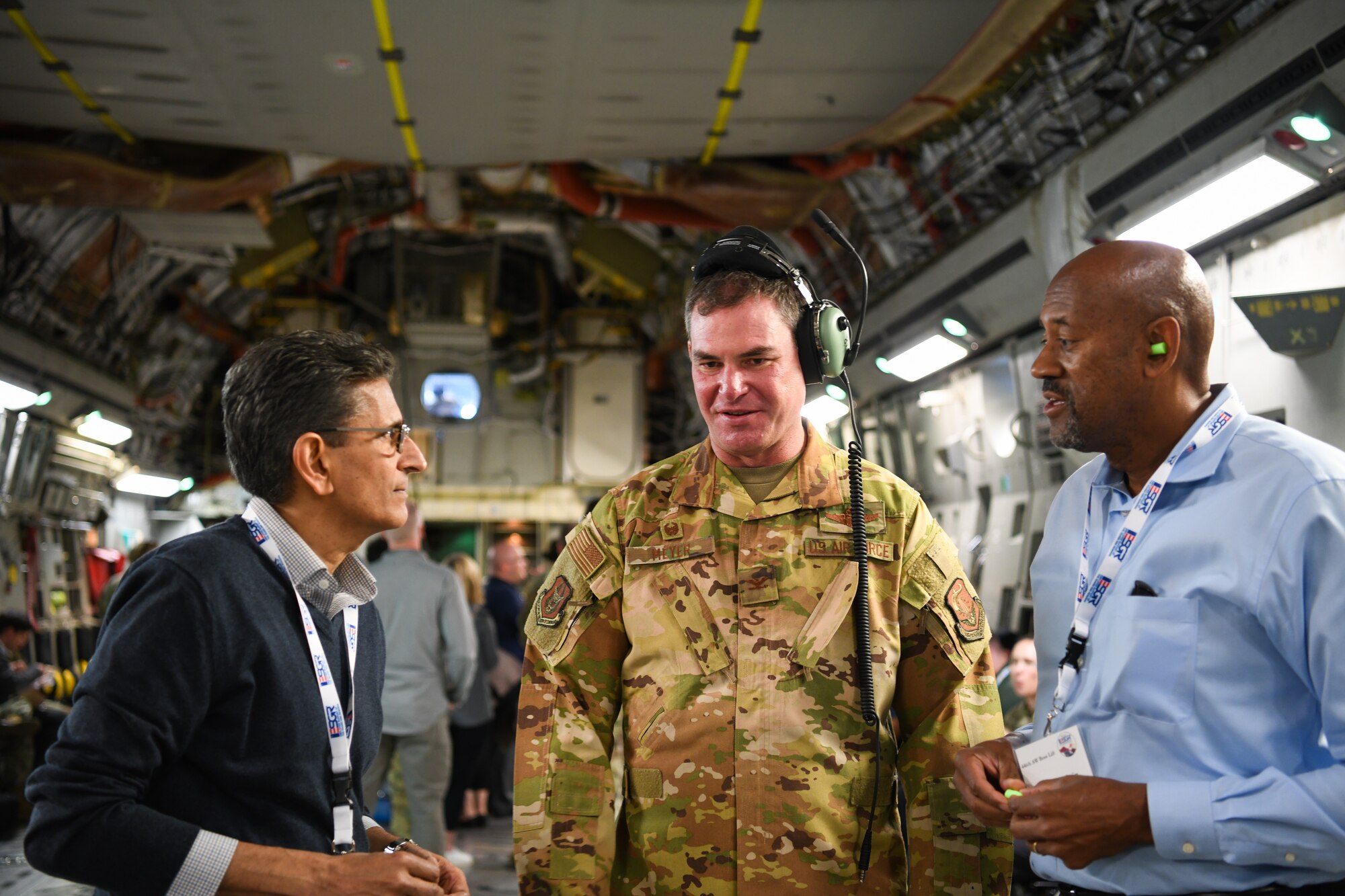 A pilot in a camo flight suit wearing a headset speaks with two civilian employers aboard a C-17 Globmeaster III in flight.