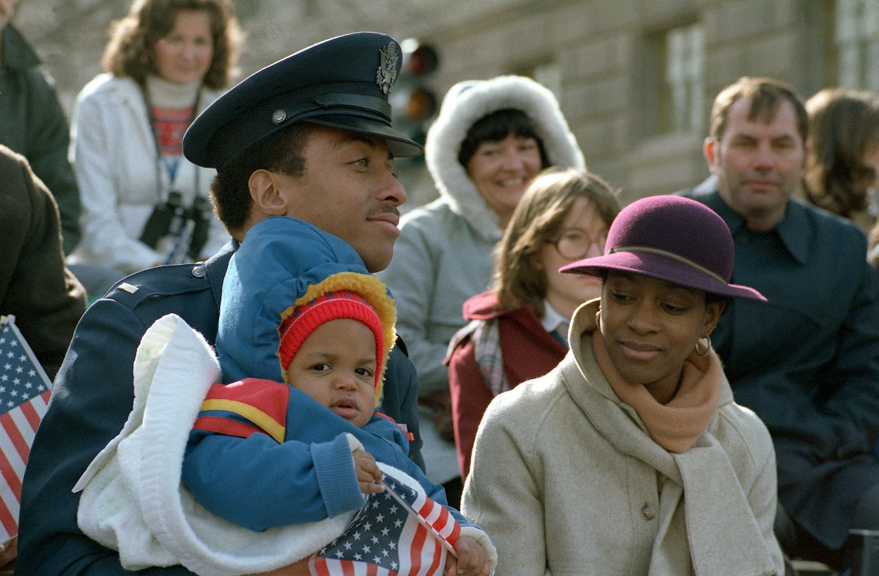 An airman holds a baby and stands next to a civilian woman in a crowd outdoors.