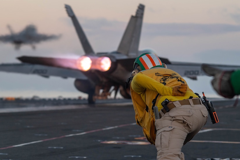 A sailor leans forward on the flight deck of a ship while signaling a military aircraft.