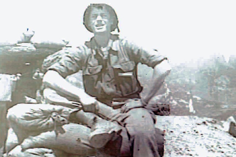 A man in uniform and helmet sits on a rock beside sandbags.