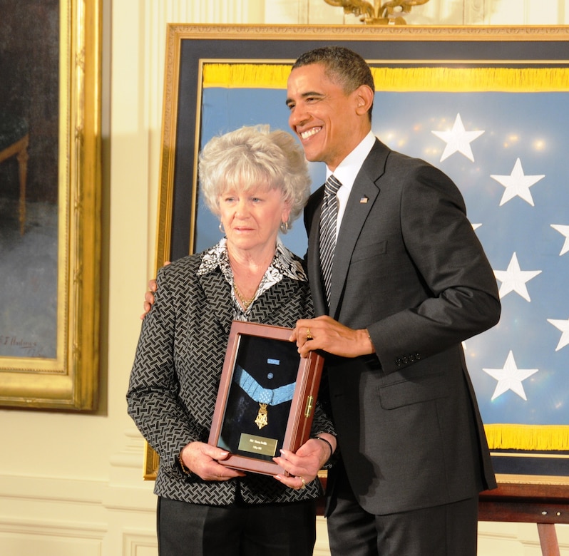 A man and a woman pose while both holding a small shadow box holding a medal.