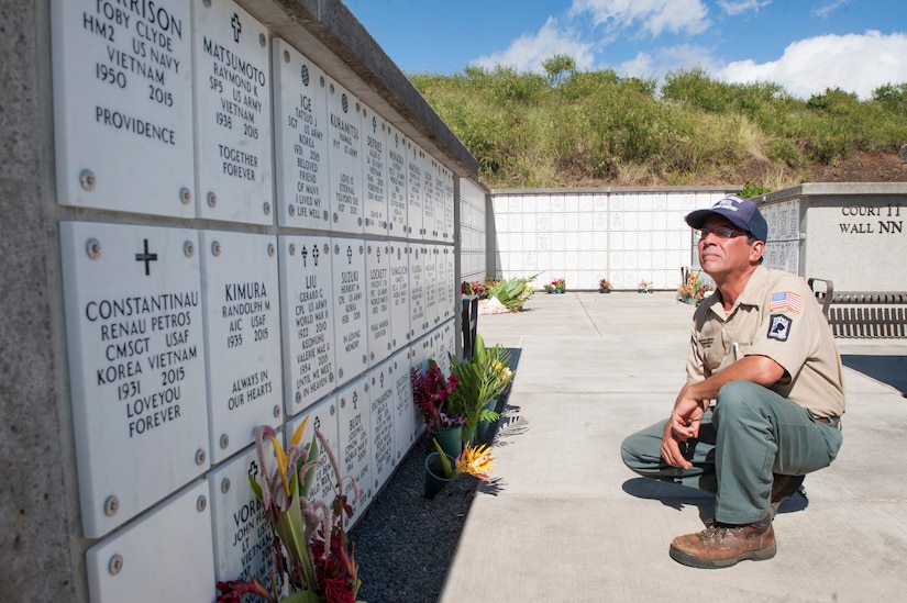 A squatting man looks at memorial plaques on a low wall.