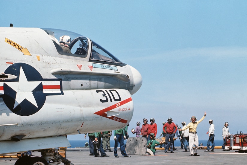 An officer signals to an aircraft parked on the deck of a Navy ship as other troops stand nearby.