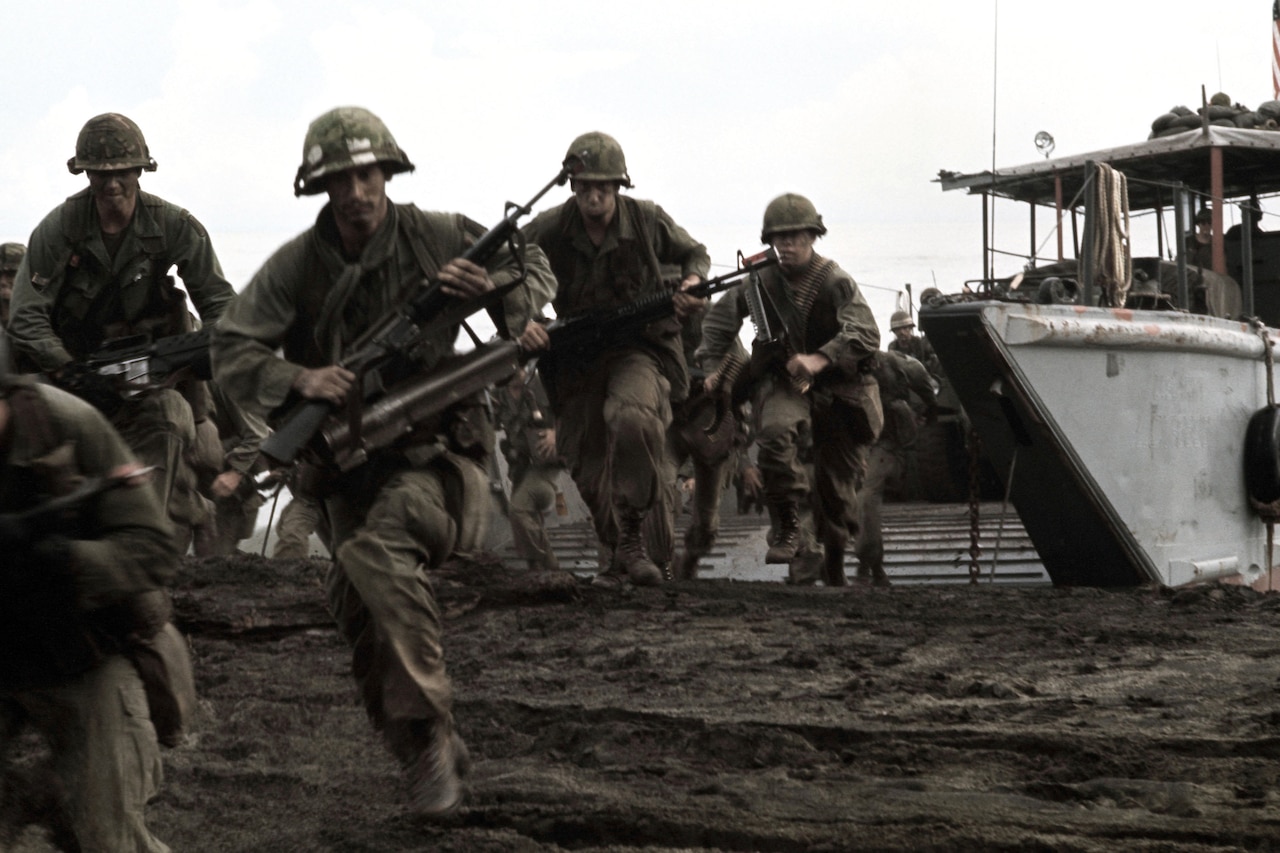 Soldiers in helmets and carrying weapons run on a beach away from a landing craft.