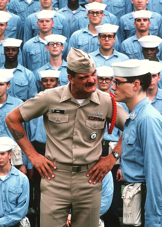 A Navy company commander, hands on hips, scowls at a recruit as others sit on bleachers in the background.