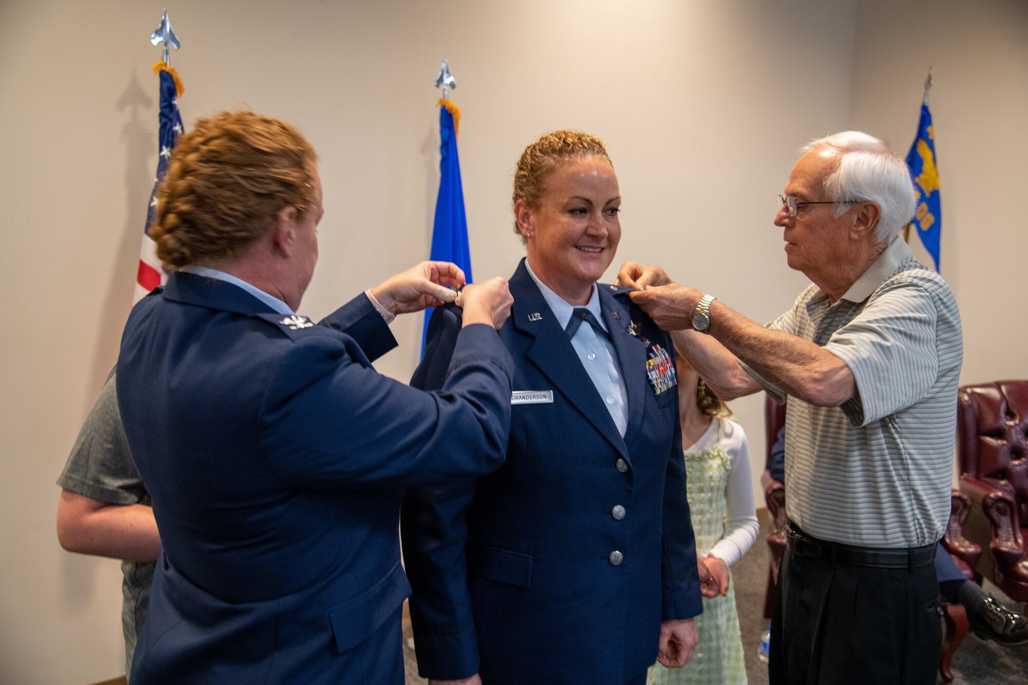 Female military member is pinned with Colonel rank by twin sister in military uniform and father in civilian clothing.