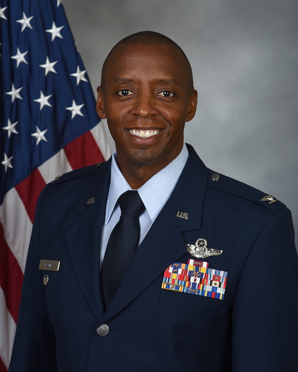 A man is seated in front of the American flag in Air Force service dress, smiling.