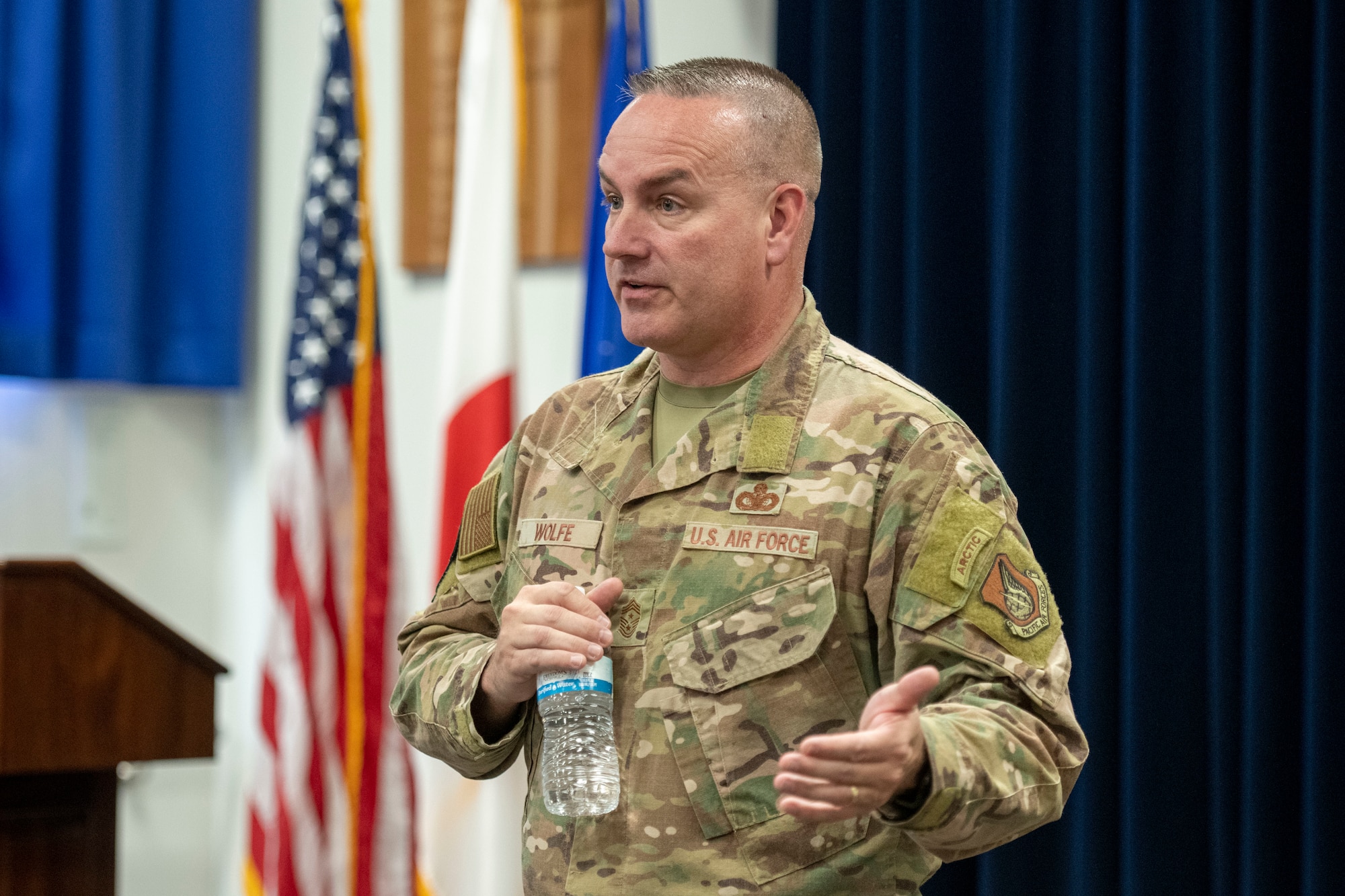 A man in a military uniform holding a water bottle speaks while gesturing