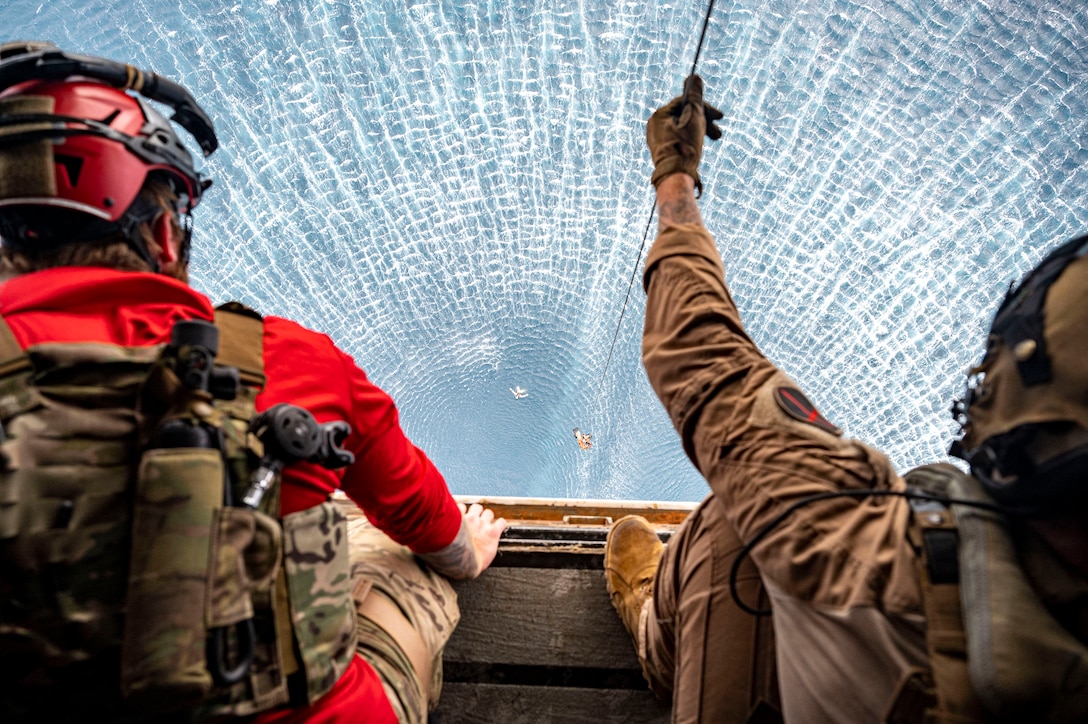 Two service members conduct hoist operations from a military aircraft flying over water.
