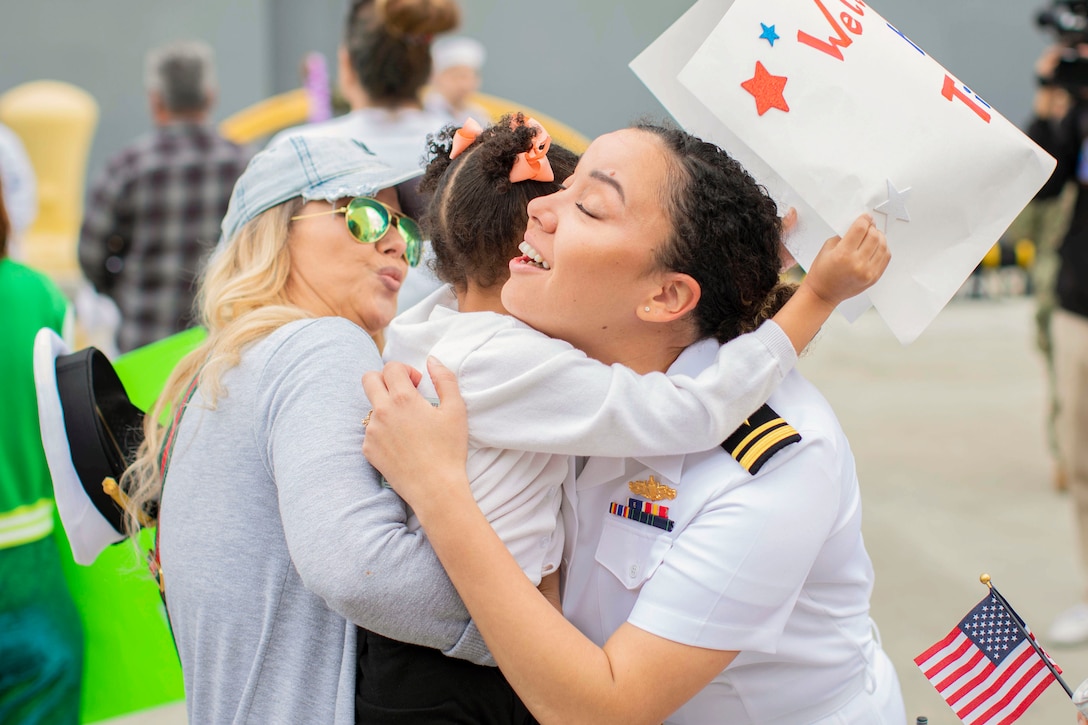 A sailor hugs two family members as people walk in the background.