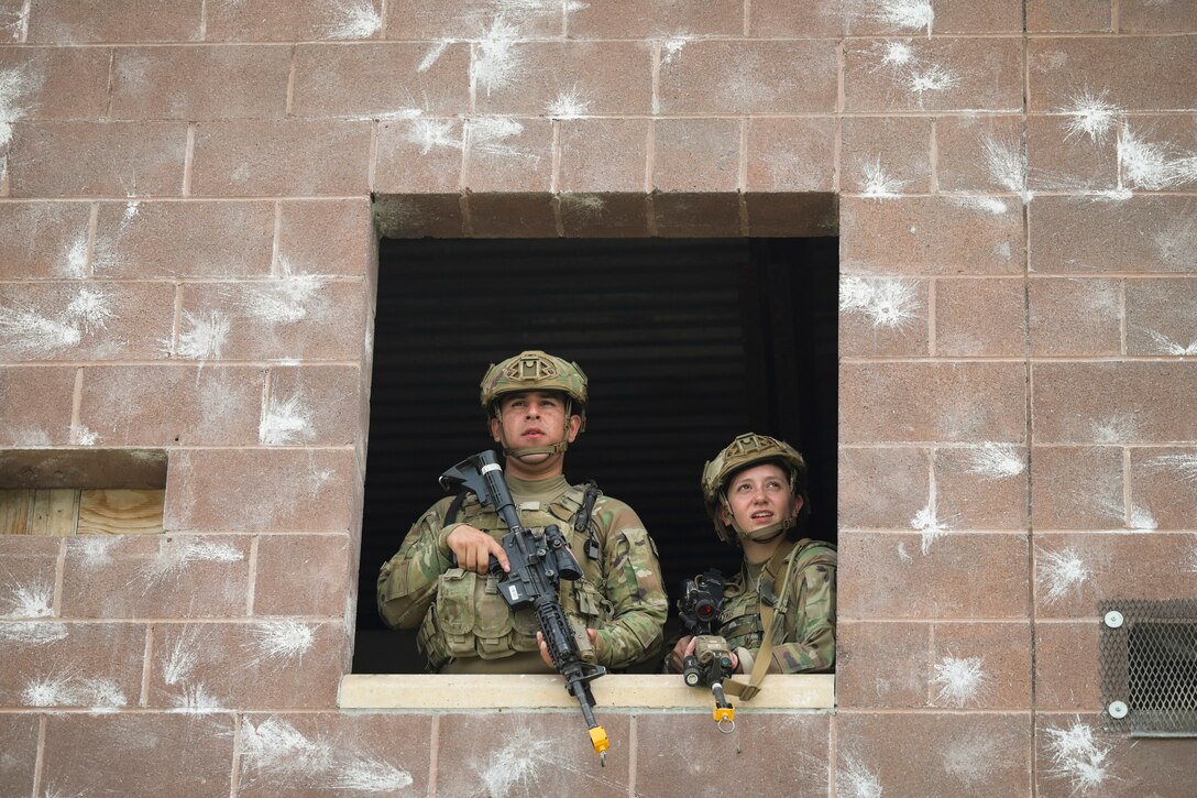 Airmen hold weapons as they look through the window of a brick wall with white markings.