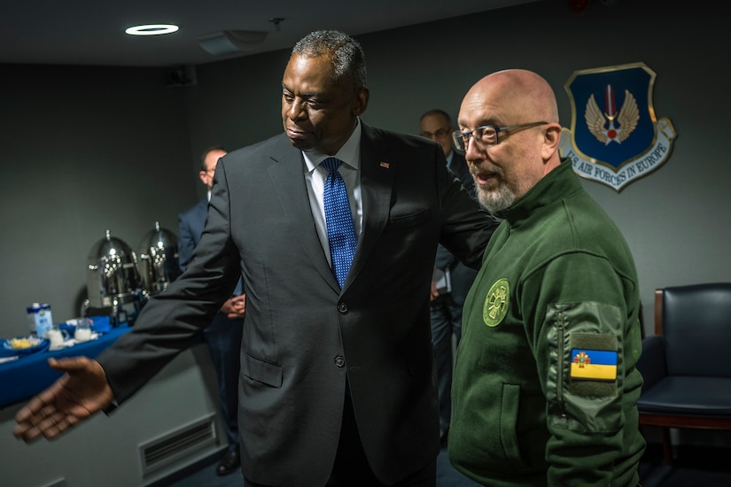 A man in a suit stands next to a man in an olive jacket with a Ukrainian flag patch on the sleeve.