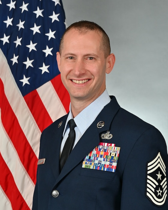 A man sits in front of an American flag, wearing U.S. Air Force service dress and smiling.