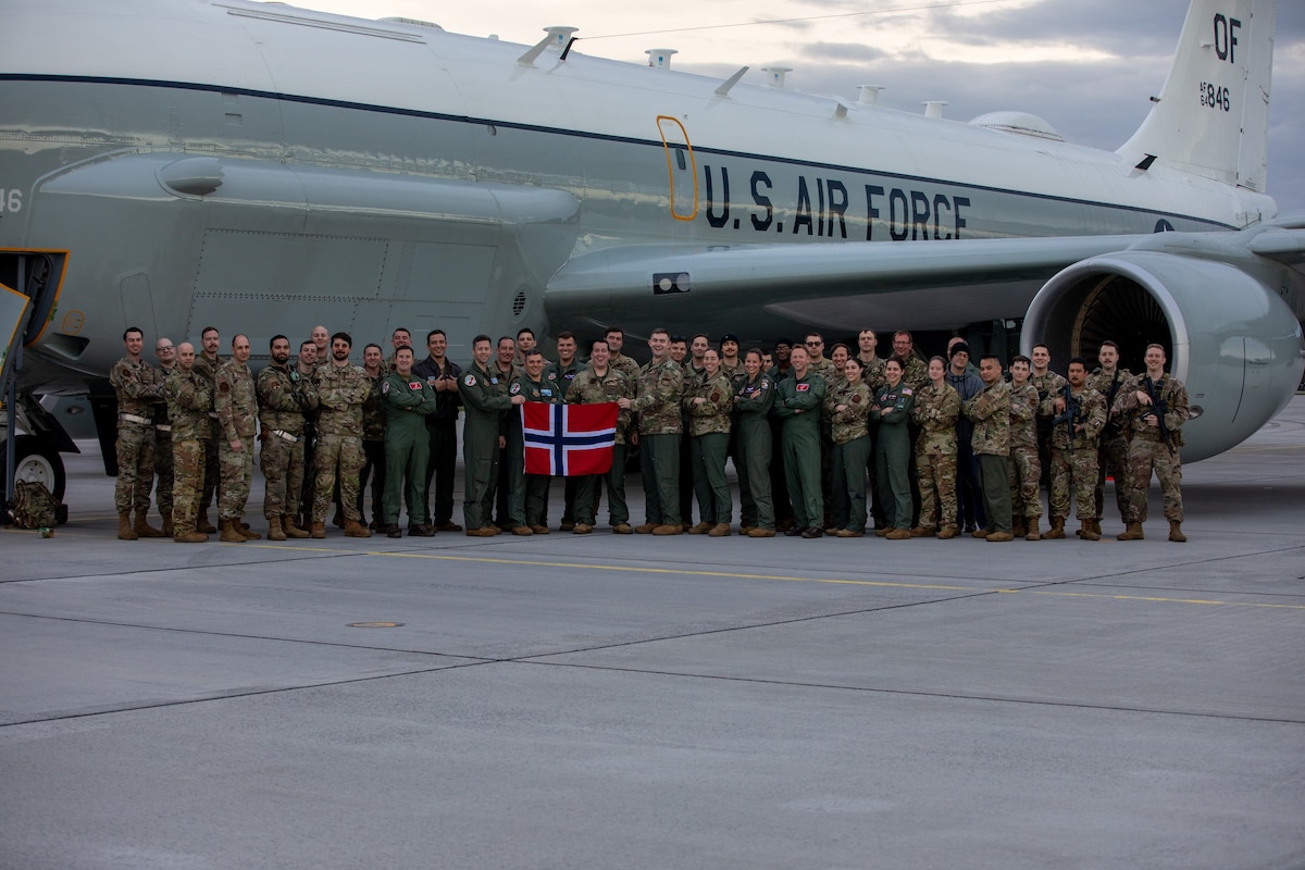 A group of military members pose in front of a U.S. Air Force aircraft with a couple of them holding a Norwegian flag