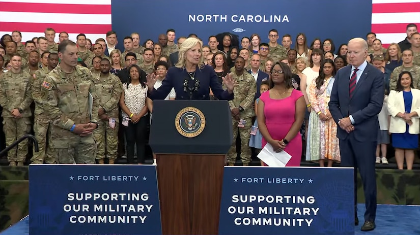 A woman stands behind a lectern. Behind her are dozens of people dressed in both military uniforms and civilian clothing. The words "Supporting our Military Community" are on signs in front of her.