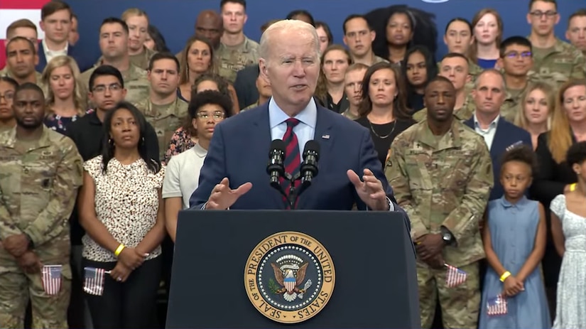 A man stands behind a lectern; behind him are dozens of people dressed in both military uniforms and civilian clothing.