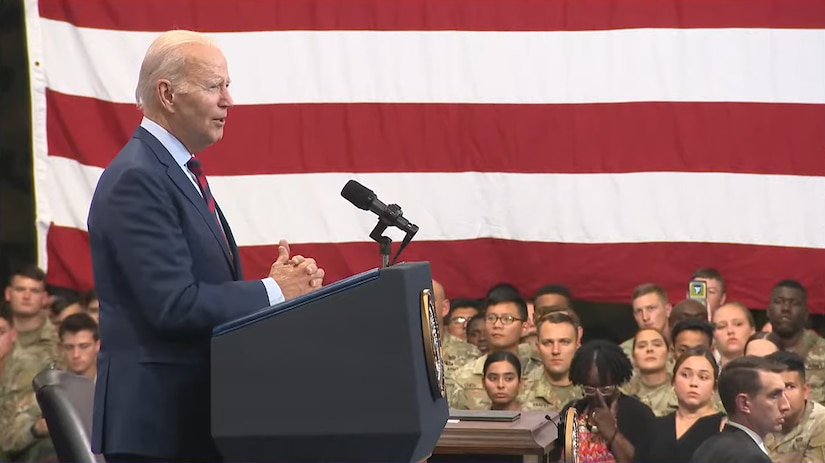 A man stands behind a lectern. Behind him are a U.S. flag and dozens of people dressed in both military uniforms and civilian clothing.