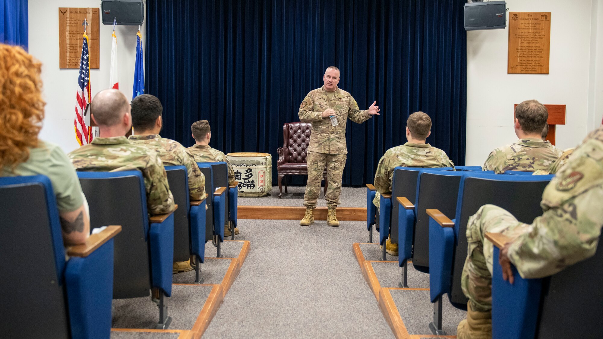 People seated in a small theater watch a man in a military uniform speak to them