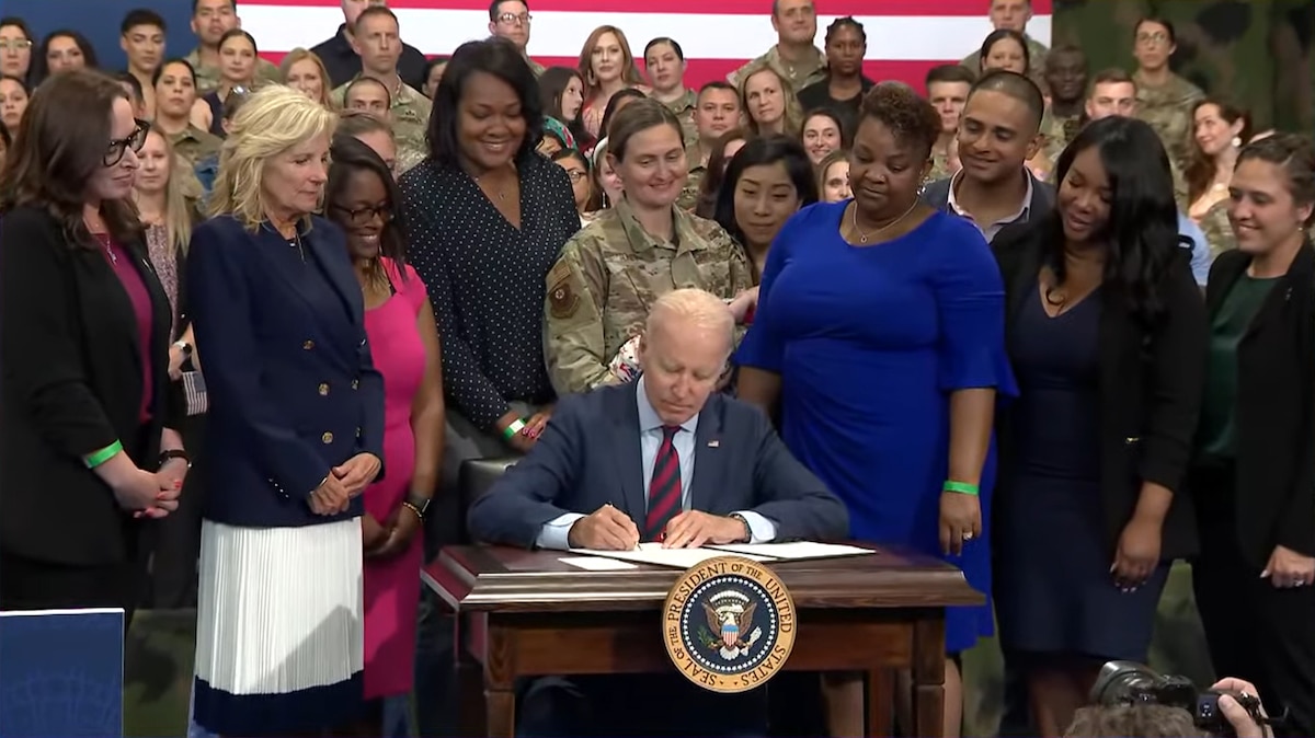 A man sits at a small table and signs a document. Behind him are a U.S. flag and dozens of people dressed in both military uniforms and civilian clothing.