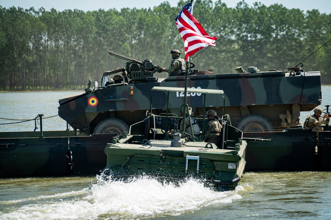 Soldiers in a U.S. boat guide Romanian soldiers in a vehicle over a temporary bridge on a river.