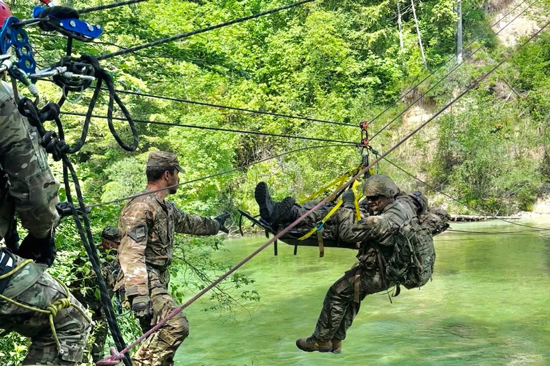 A soldier on a rope pulls another service member across a river as a third waits.