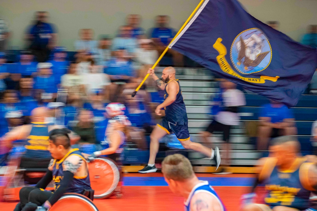 A service member runs with a flag as others move through a basketball court in wheelchairs.