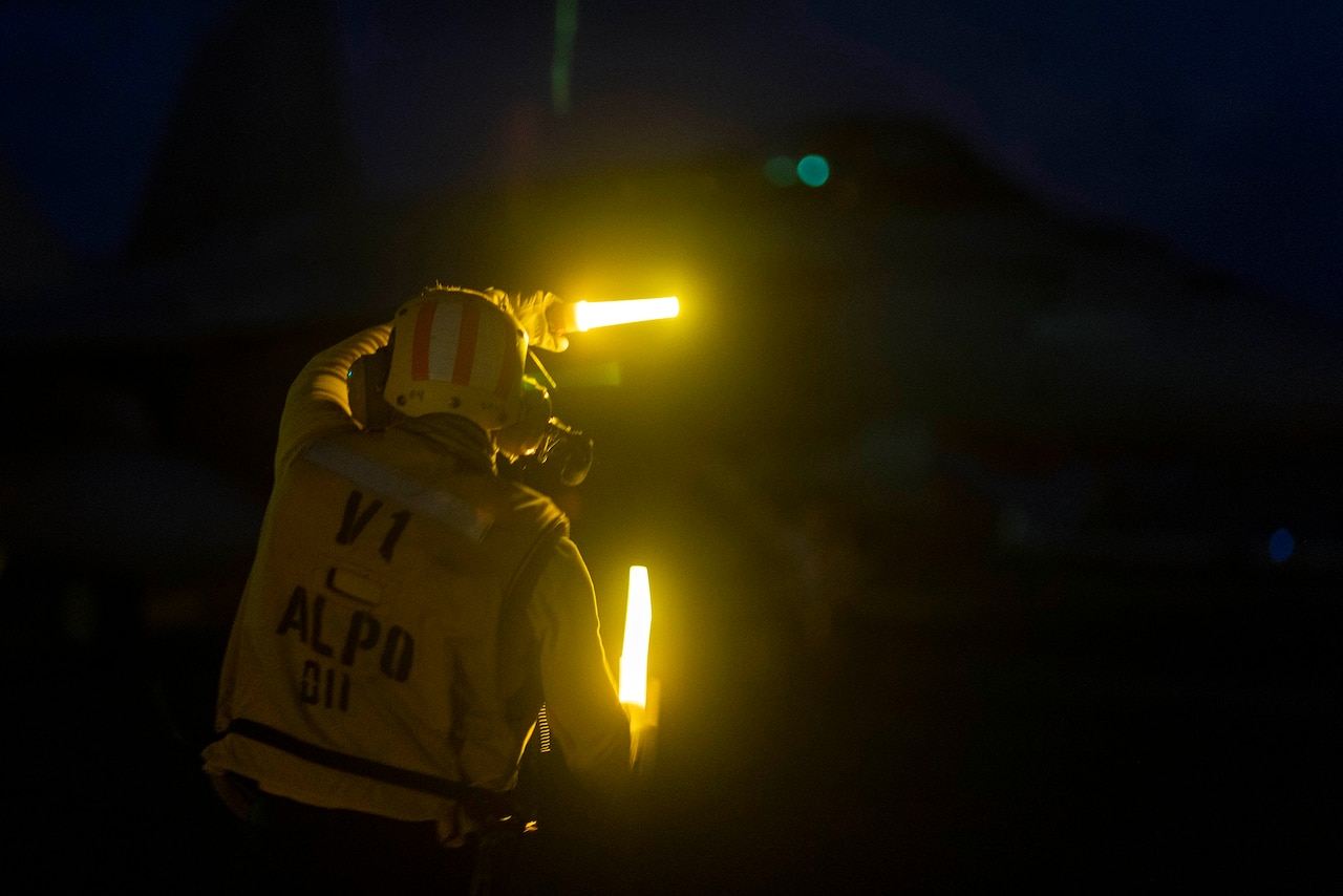A sailor waves lights to direct a military aircraft on the flight deck of a ship at night.