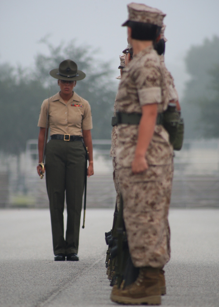 A Marine Corps staff sergeant stands and looks at recruits standing in a line outdoors.