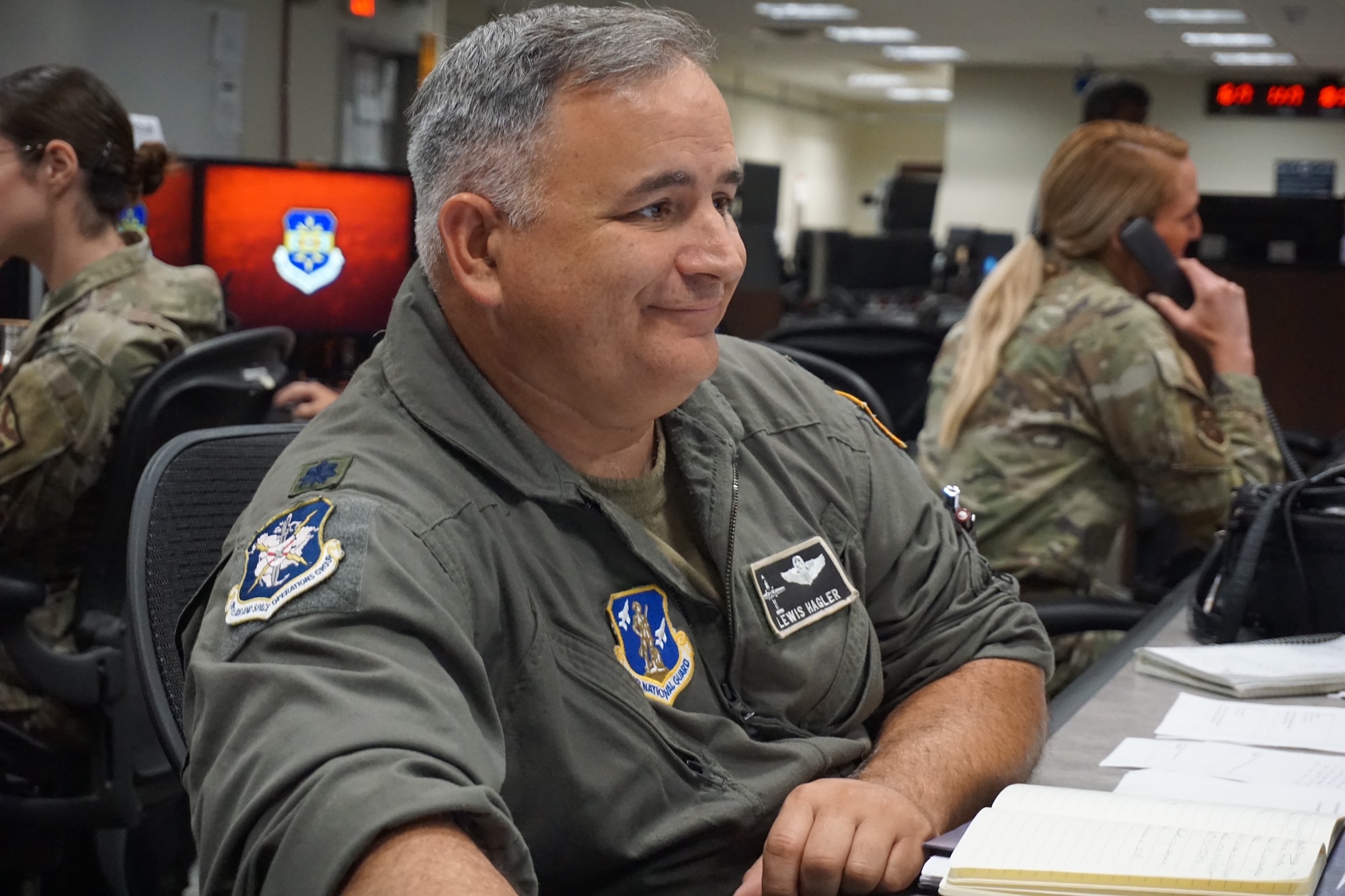 photo of a uniformed U.S. ANG Airman sitting at table filled with computers with other U.S. Air Force Airmen working in the background