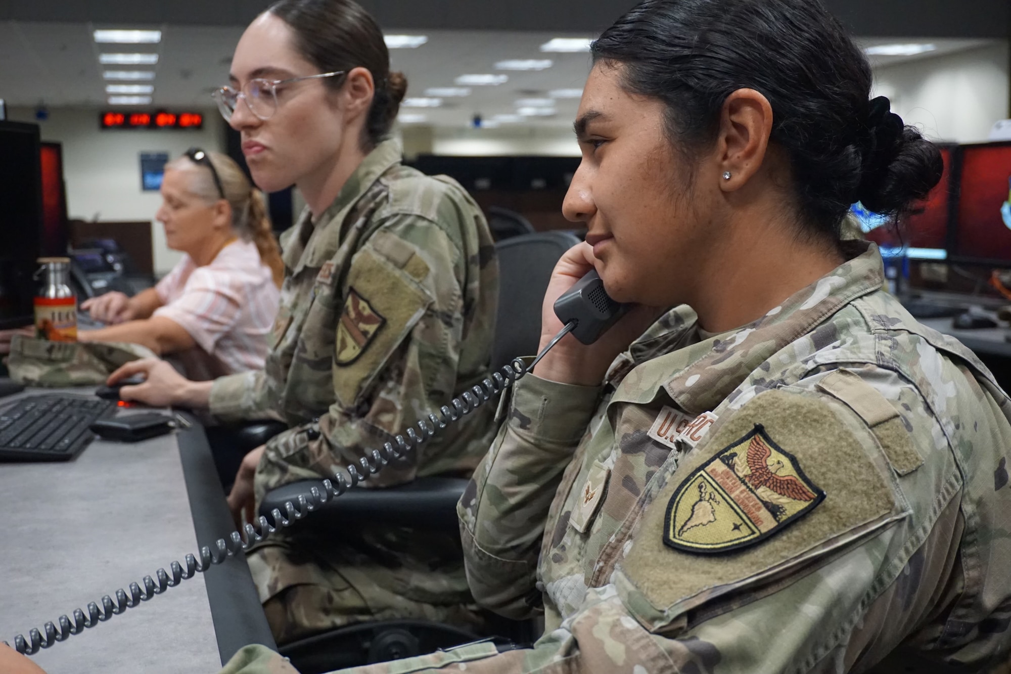 photo of two uniformed U.S. Air Force Airmen and one civilian sitting at computers, one Airman is holding a phone