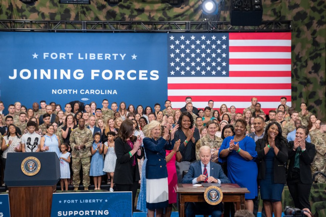 President Joe Biden sits at a desk flanked by a standing, applauding crowd, including the first lady.