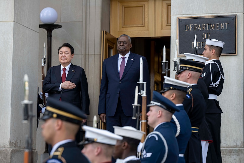 Two men wearing suits stand atop steps overlooking a military ceremonial guard.