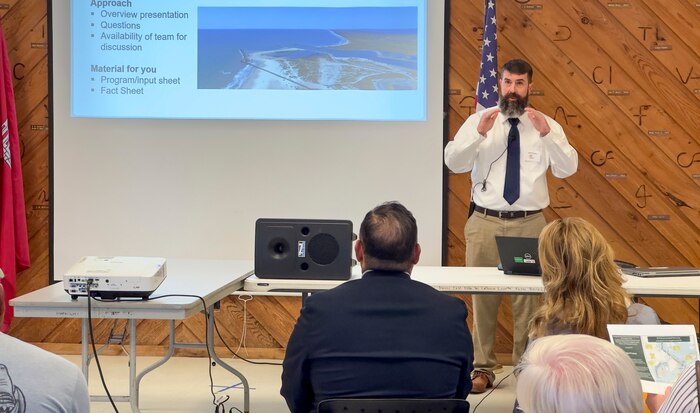 a man is standing in front of a crowd, next toa projection screen, talking