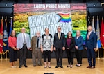 Eight people stand on a wood-floor stage in front of a large rainbow-colored screen displaying the words, “LGBTQ Pride Month. Being Your Authentic Self.”