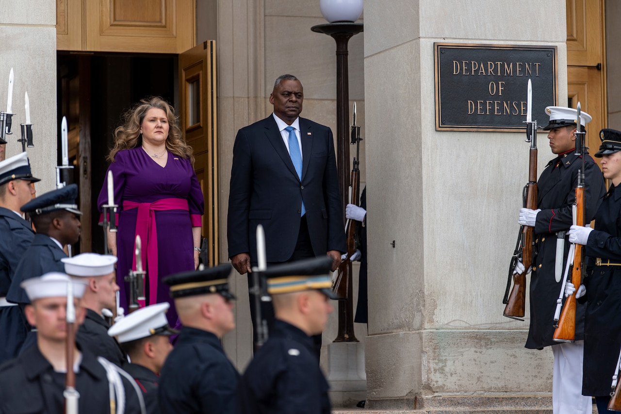 A man and woman stand atop a staircase overlooking a military ceremonial guard.
