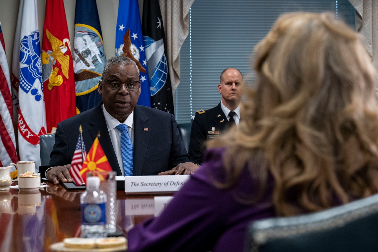 A man in a suit addresses a woman from across a table at which both are seated.