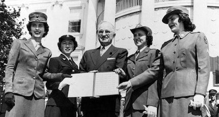 A man stands outside. He is flanked by women in military uniforms on both sides. He holds a document in outstretched hands.